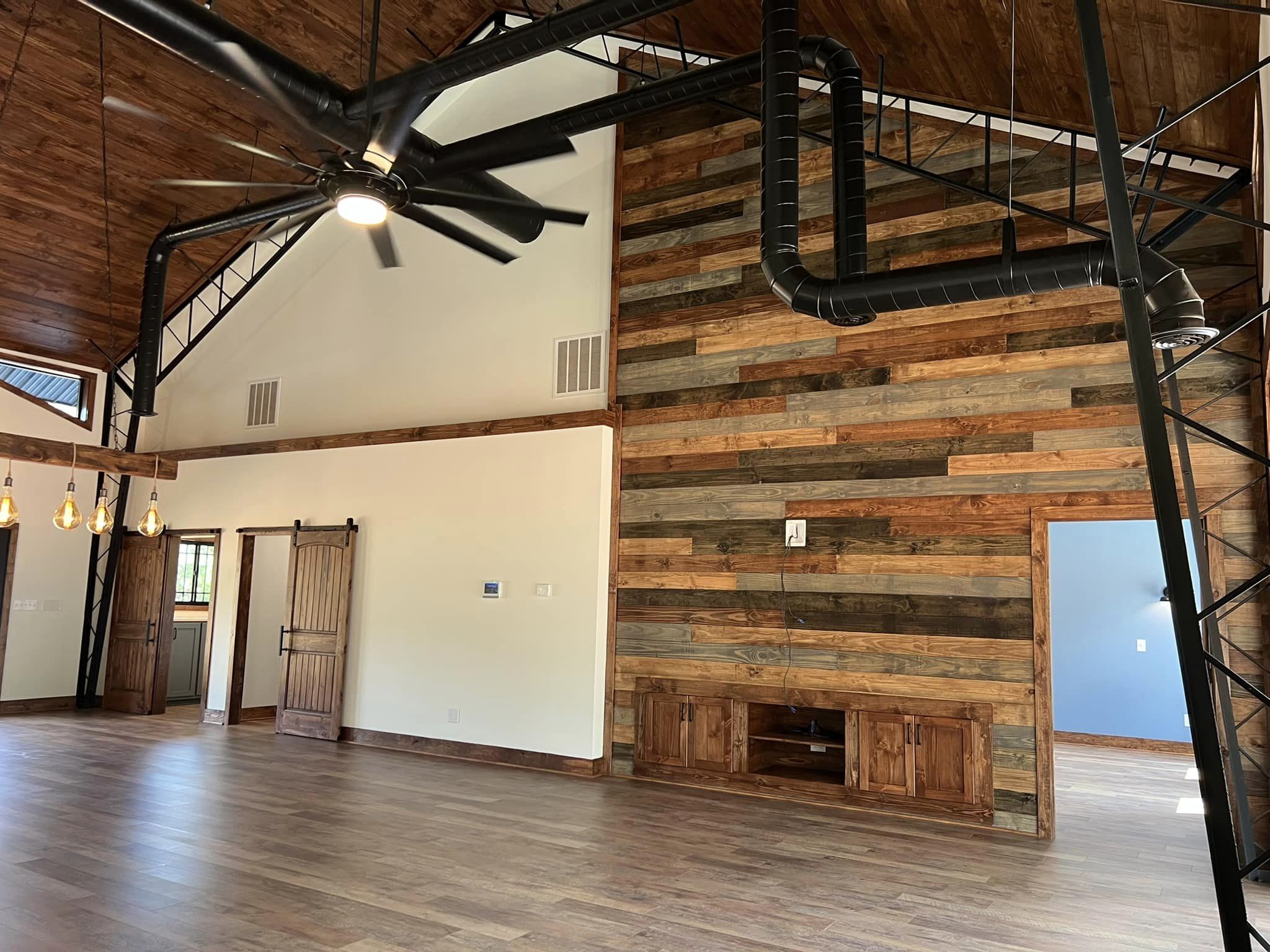 A spacious room with wooden floors and walls, echoing a modern Barndominium style, featuring a large ceiling fan and industrial black ductwork. A sliding barn door graces one wall, adorned with hanging pendant lights. An alcove with shelves adds charm to this Evermore-inspired space.
