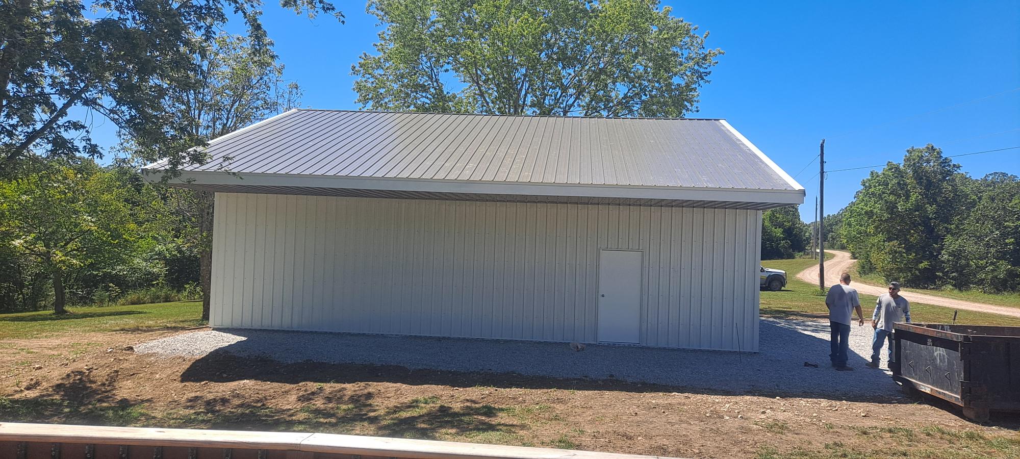 A large, newly constructed metal garage with a gabled roof stands on a gravel-covered area in Missouri. Two people are nearby, and trees and a clear blue sky enhance the scenic backdrop.
