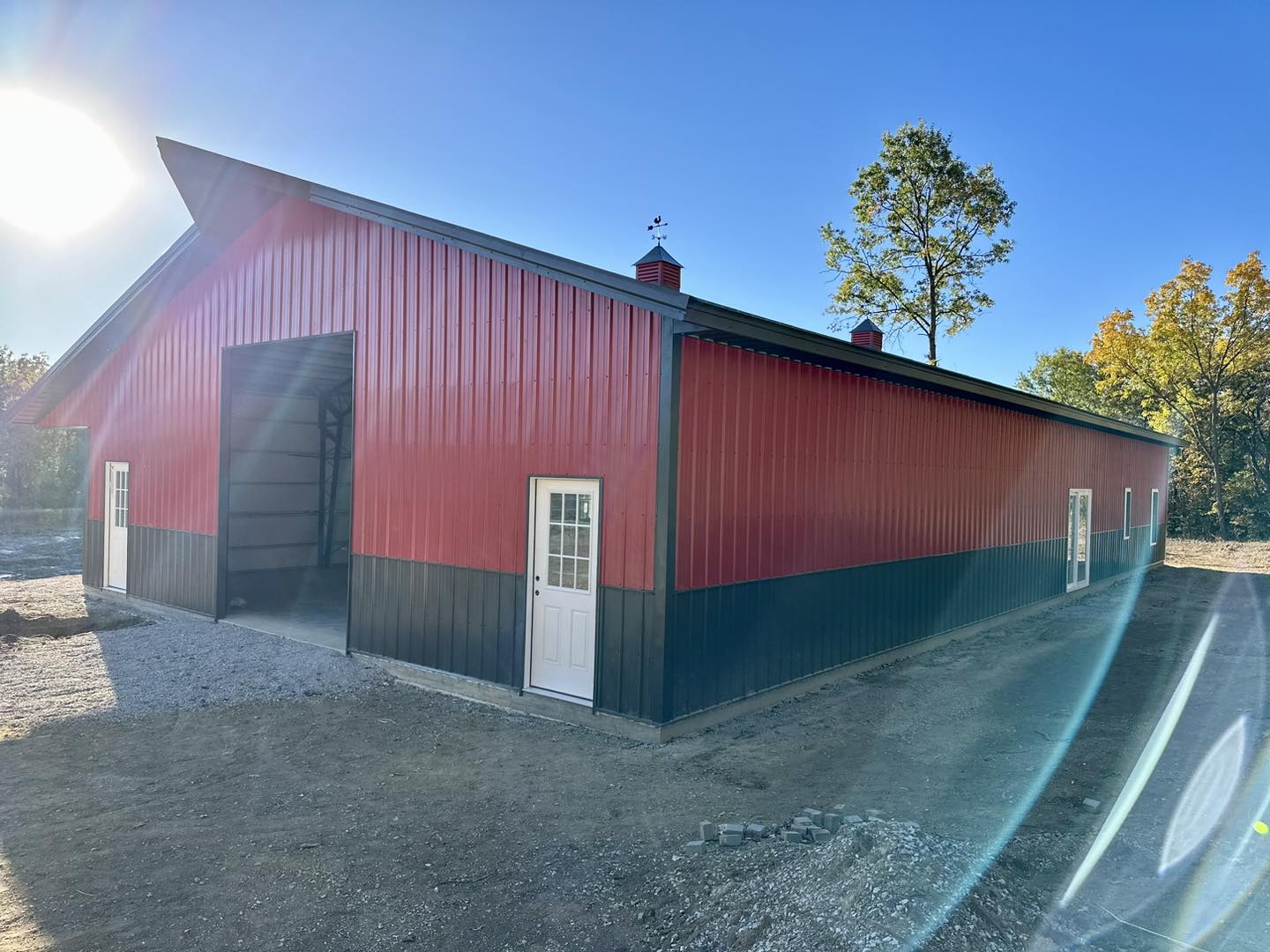 A large red and black barn with white doors, marked as DeLuca Farms, stands under a clear blue sky. The sun shines brightly in the top left corner, while a few trees with green leaves add charm to the background. The ground around this barndo is covered in gravel.