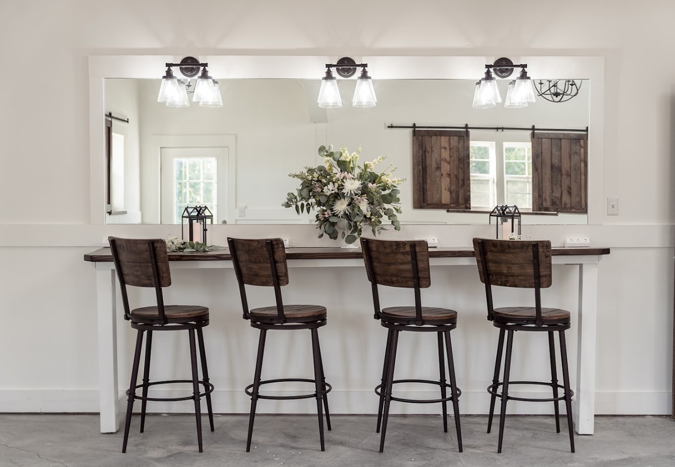 A modern kitchen bar setup at Stone Briar Farms features four wooden stools facing a large mirror. The counter boasts a floral arrangement, and three pendant lights hang above. This elegant event venue showcases white walls and rustic wooden shutters on the windows.