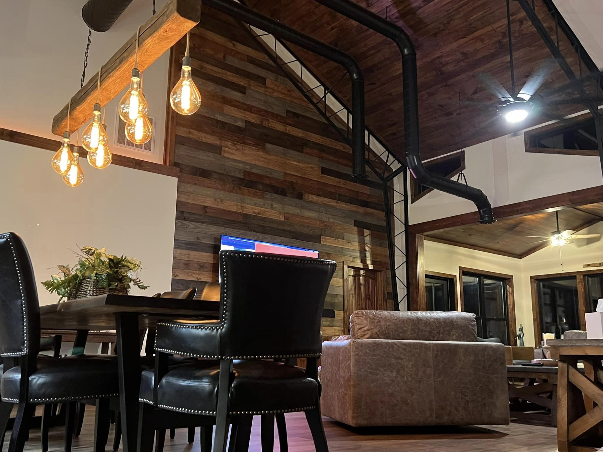 A cozy living room in the Evermore Barndominium features a rustic wooden ceiling and wall. A dining table with black leather chairs sits under warm, hanging light bulbs. A beige sofa faces a TV, while large windows frame the background beautifully.