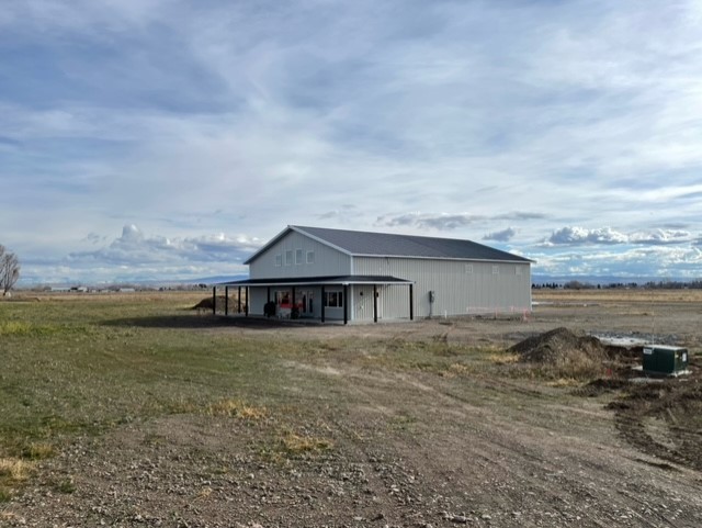 A large, gray barndominium with a metal roof stands in a vast Idaho field under a cloudy sky. The ground is a mix of grass and dirt, with some equipment near the structure. Sparse trees and distant mountains are visible in the background, adding to the rustic design.