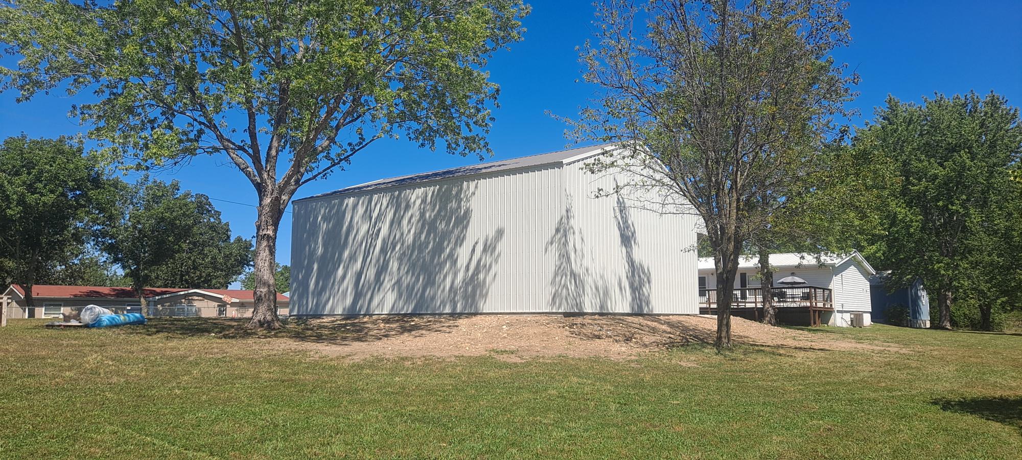A large gray metal barn, reminiscent of impressive Metal Garage Buildings found in Missouri, stands on a grassy hill flanked by trees with shadows cast on its surface. A small white structure is partially visible to the right under the clear blue sky.