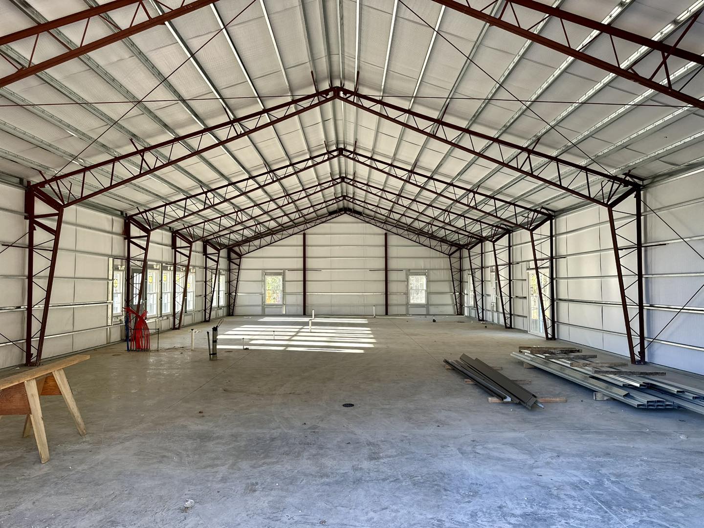 Interior of an unfinished warehouse with exposed metal beams and a concrete floor, reminiscent of a rustic barndo style. Sunlight filters through open windows, casting shadows over scattered building materials and a wooden sawhorse on the floor, evoking the charm of DeLuca Farms.