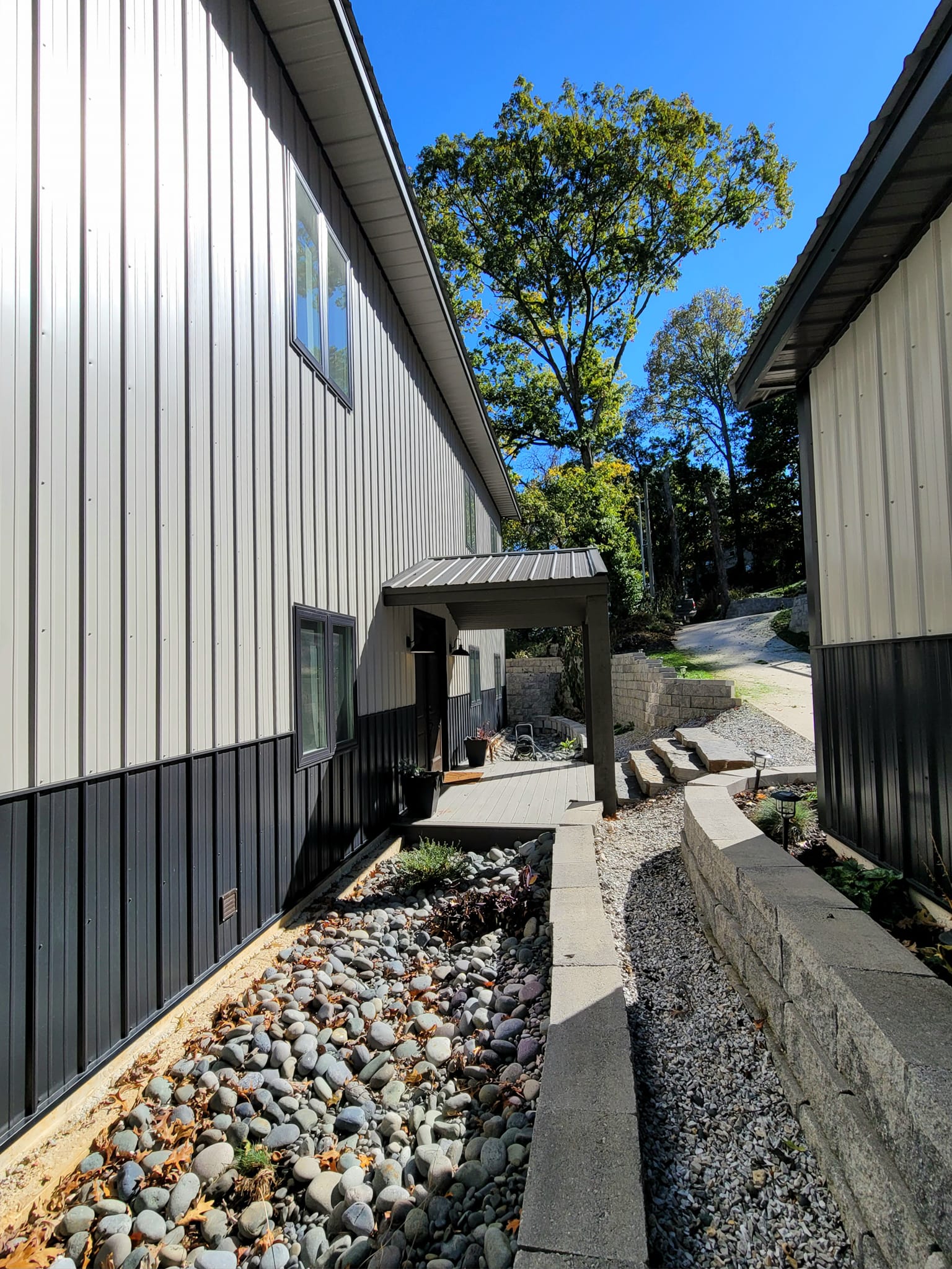 Side view of a modern building with a metal facade, reminiscent of the sleek design seen at Table Rock. It features a covered entrance and a landscaped area with stones and a retaining wall lining the walkway. Trees and a bright blue sky provide a stunning backdrop.