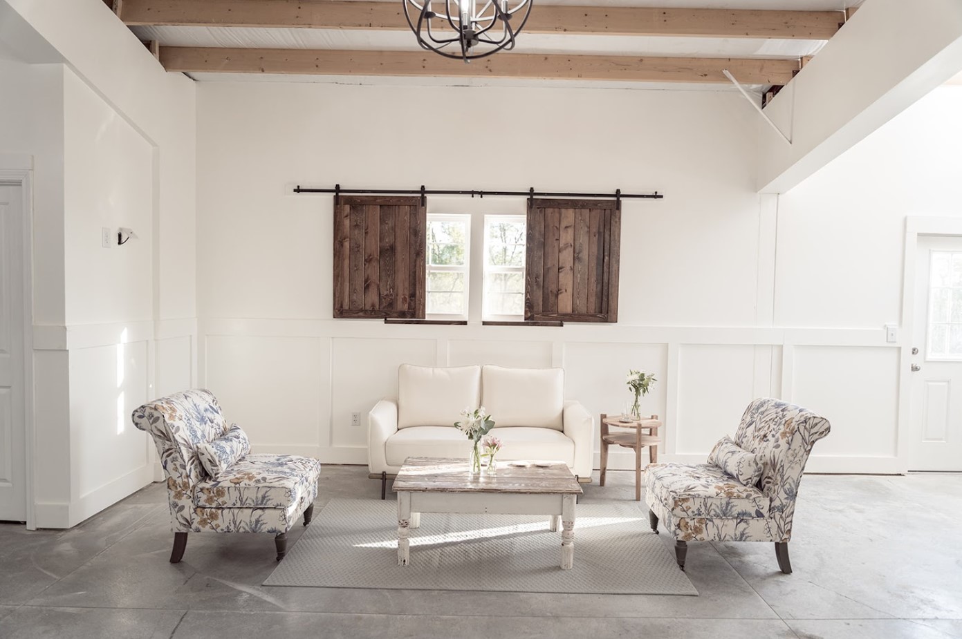 A bright, minimalist living room at Stone Briar Farms features a white sofa and two floral-patterned armchairs. A wooden coffee table sits in front of the sofa, beneath exposed beams and a black chandelier. Rustic wooden window shutters add charm, perfect for an intimate event venue.