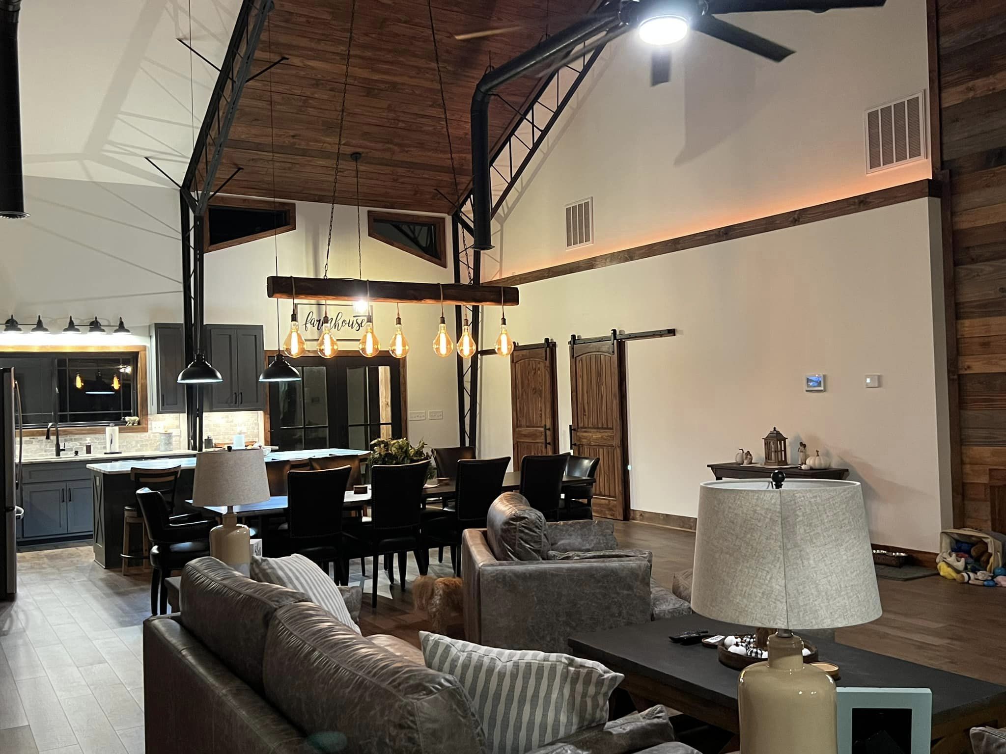 A spacious, modern living room in the Evermore Barndominium boasts high ceilings and exposed beams. Features include a dining area with a hanging light fixture, a kitchen with dark cabinets, a cozy seating area, and rustic wooden accents such as sliding barn doors.