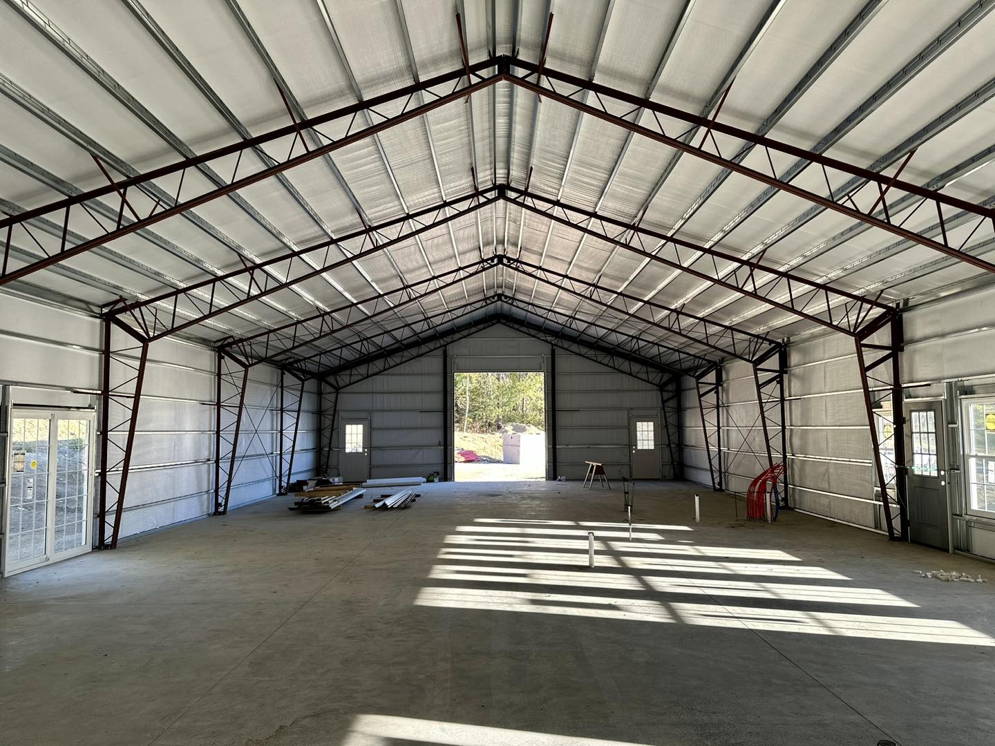 The interior of the expansive metal warehouse at DeLuca Farms boasts high ceilings and steel beams. Sunlight floods through open doors, weaving shadows on the concrete floor. A few construction materials lie scattered across this versatile barndo space.