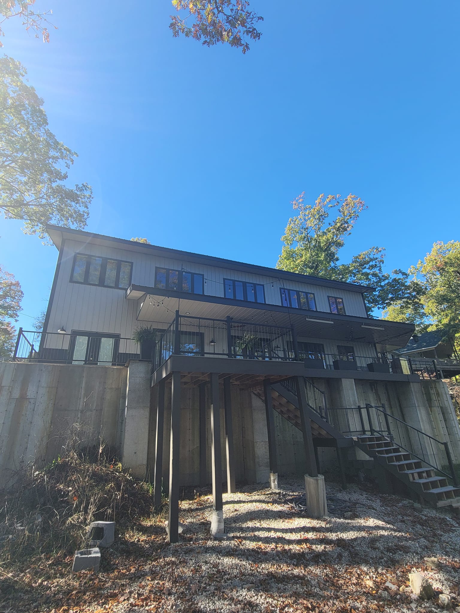 A modern two-story Lakehouse on stilts features a striking black and white exterior. Elevated with a staircase leading up, it is surrounded by trees and set against a clear blue sky on a sunny day, reminiscent of the serene beauty of Table Rock.