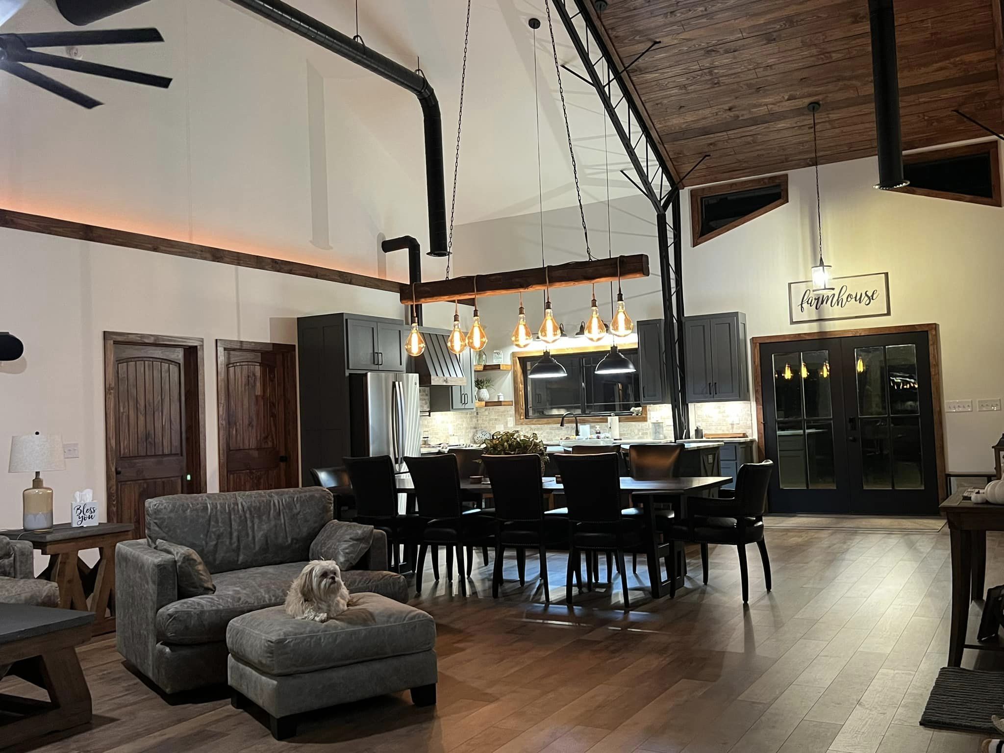 A cozy living space in a barndominium featuring a modern kitchen with dark cabinets, a large dining table, and rustic decor. A dog lounges on a gray cushioned sofa. Warm lighting and wooden accents complete the welcoming atmosphere.