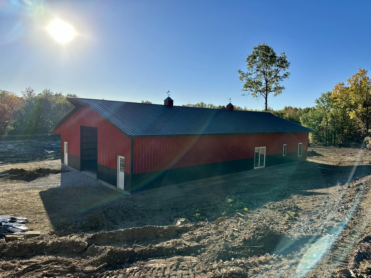 Under the clear blue sky, a large red barndo with a black roof stands prominently. Surrounded by a dirt area and trees in the background on DeLuca Farms, the sun shines brightly, casting warm rays across the picturesque scene.