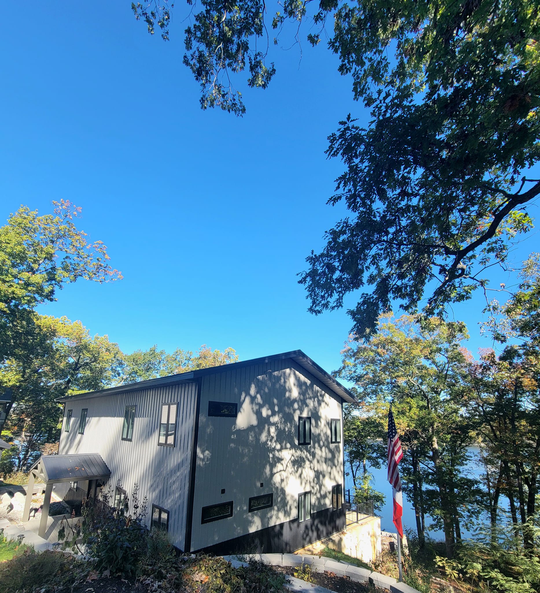 A modern, two-story lakehouse with a light gray exterior is nestled among trees. An American flag proudly waves in the yard. In the background, a clear blue sky and glimpses of Table Rock through the trees add to its charm.