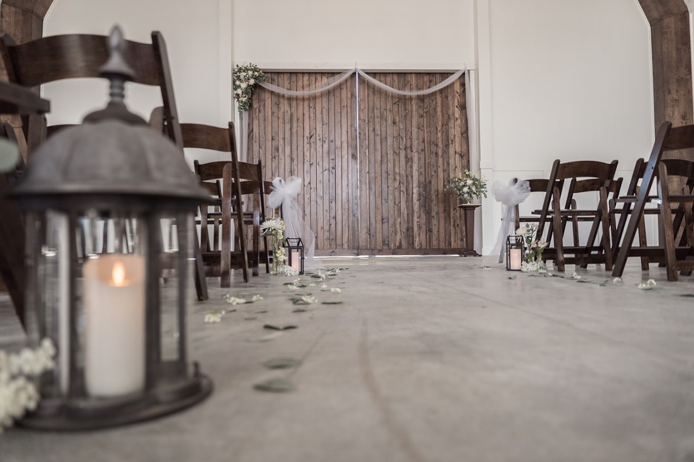 A rustic wedding ceremony setup at Stone Briar Farms features wooden chairs lining an aisle adorned with white flowers and lanterns. The entrance boasts large wooden double doors draped in white fabric, capturing the essence of this idyllic event venue.
