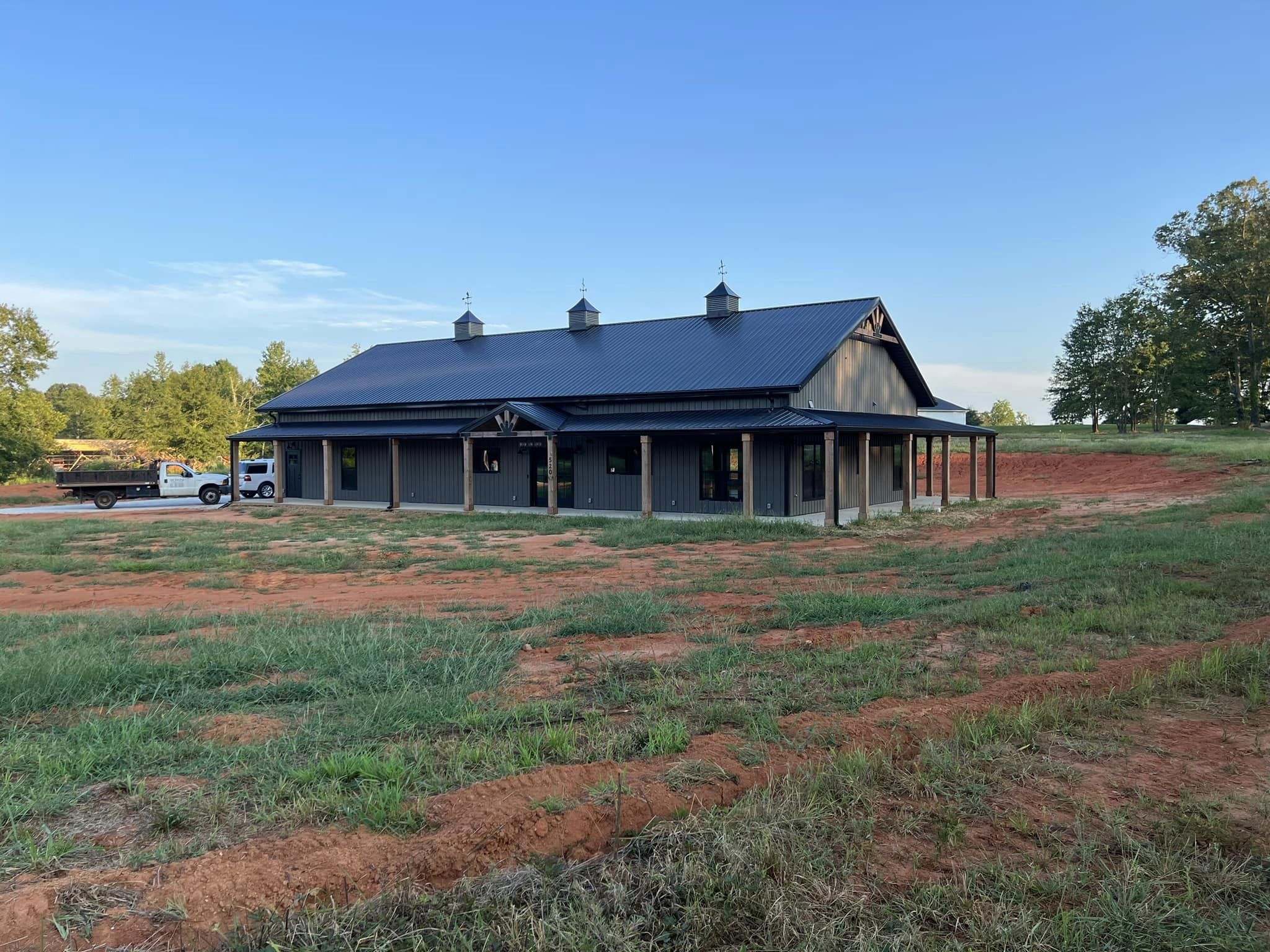A newly constructed barndominium with a dark metal roof sits in a grassy field under a clear blue sky, embodying the Evermore charm. Several trees and a parked truck are visible in the background.