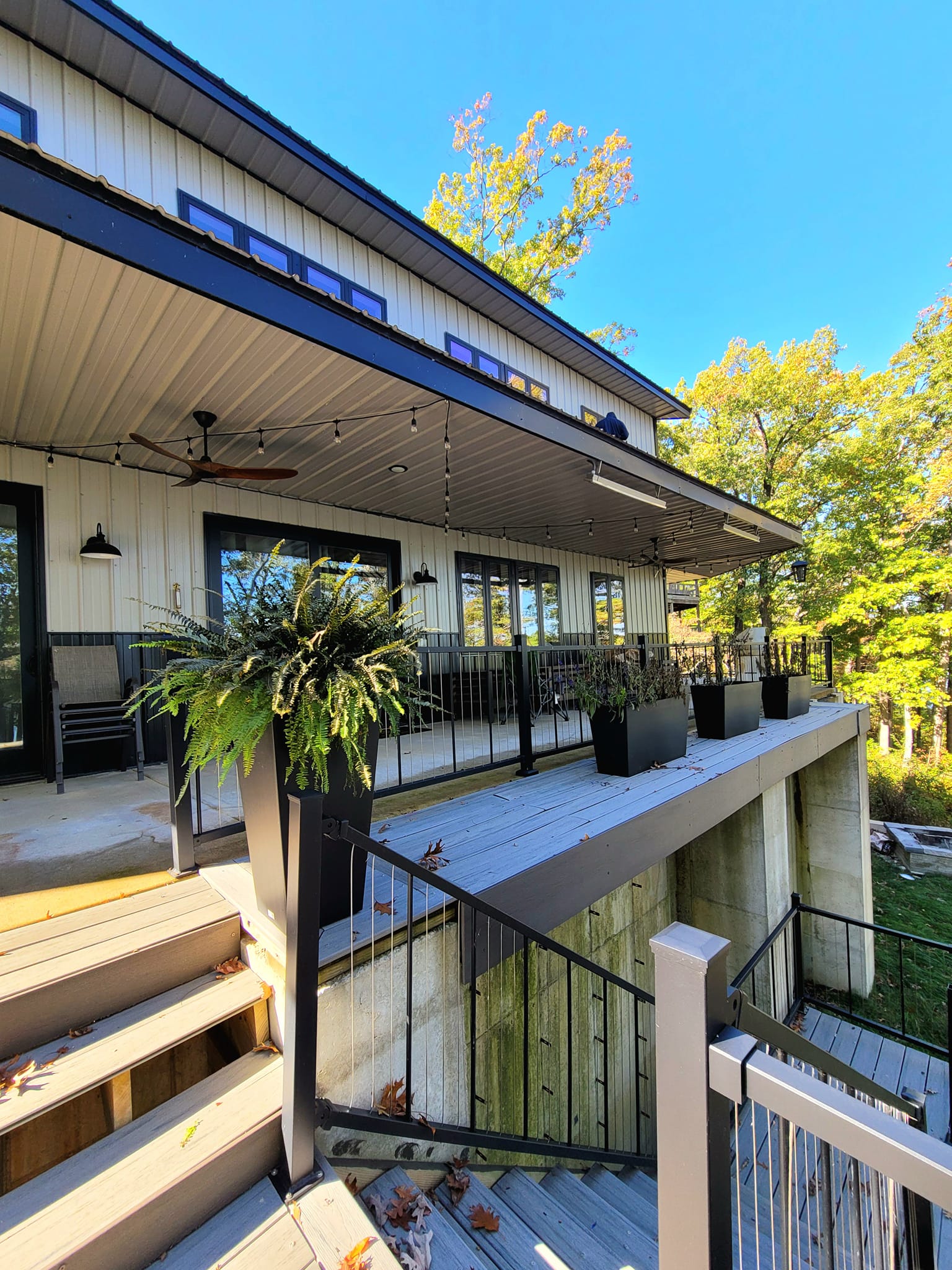The modern Table Rock house exterior boasts a large covered porch adorned with string lights. Black railings and potted plants enhance the charm, while wooden steps descend to a lower level. Framed by trees, the picturesque setting under a clear blue sky completes this inviting Red Iron Lakehouse oasis.