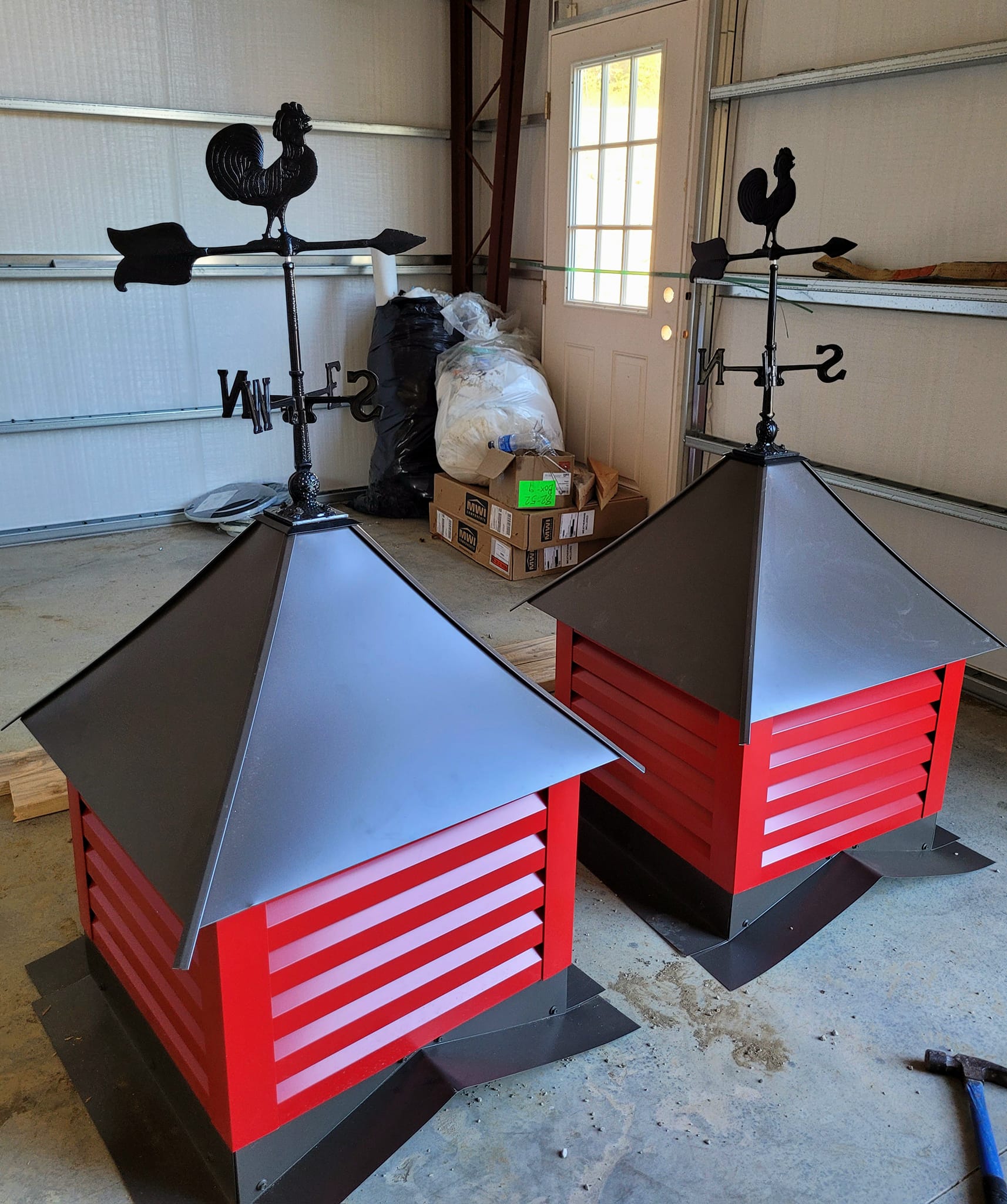 Two red and black roof cupolas with rooster weathervanes from DeLuca Farms sit on a garage floor. One is slightly in front of the other, capturing the rustic charm of a barndo aesthetic. Cardboard boxes and a closed door are in the background.