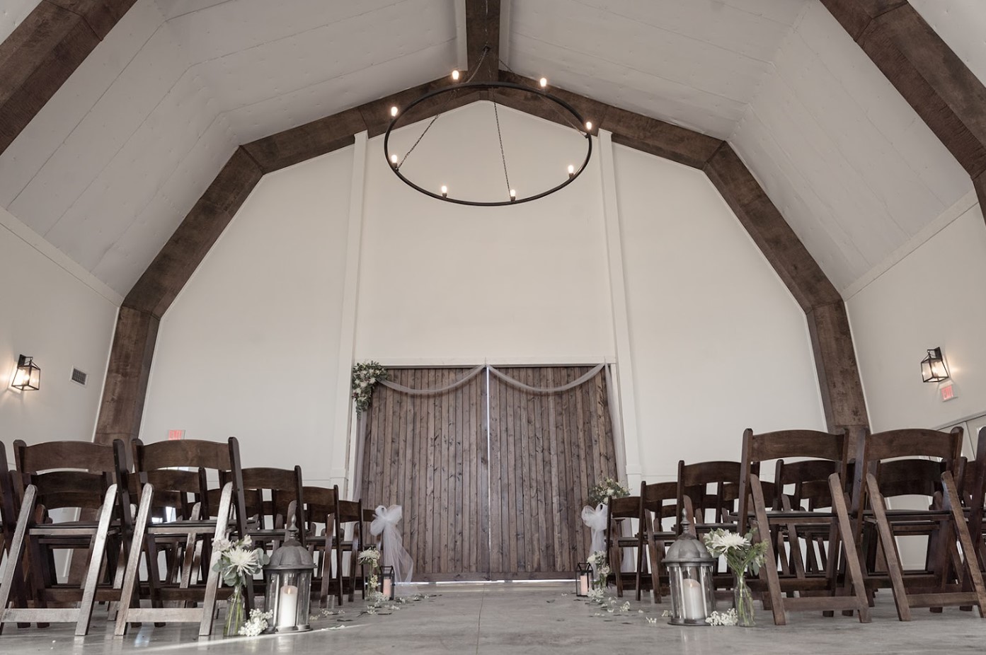 Stone Briar Farms offers a rustic wedding chapel adorned with wooden beams and a grand chandelier. As an enchanting event venue, it features dark wooden chairs in rows along the aisle, illuminated by lanterns and flowers, leading to a large wooden double door at the front.