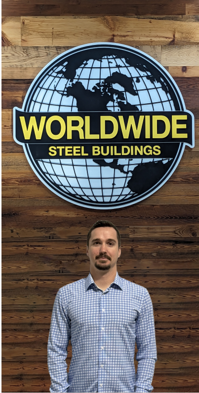 A man with glasses and a mustache is sitting at a desk cluttered with paperwork and a computer. He is wearing a beige polo shirt with a logo featuring the word "Builder." The background shows office supplies, a small framed photo, and colorful test buttons scattered around.