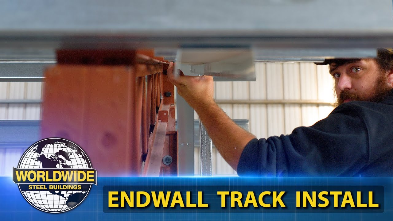 A worker meticulously installs an endwall track inside a metal building, carefully positioning the component. The image showcases a logo reading "Worldwide Steel Buildings" with text: "Endwall Track Install." This scene captures both skill and precision characteristic of expert installations.