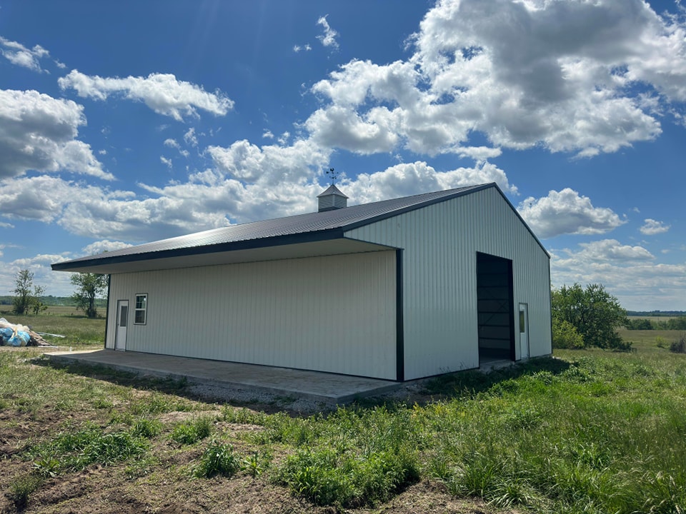 A large, white, metal barn with a black roof stands in an open grassy area under a bright blue sky with scattered clouds. The barn has a small weather vane on top and several windows and doors. The surrounding landscape is green and peaceful.
