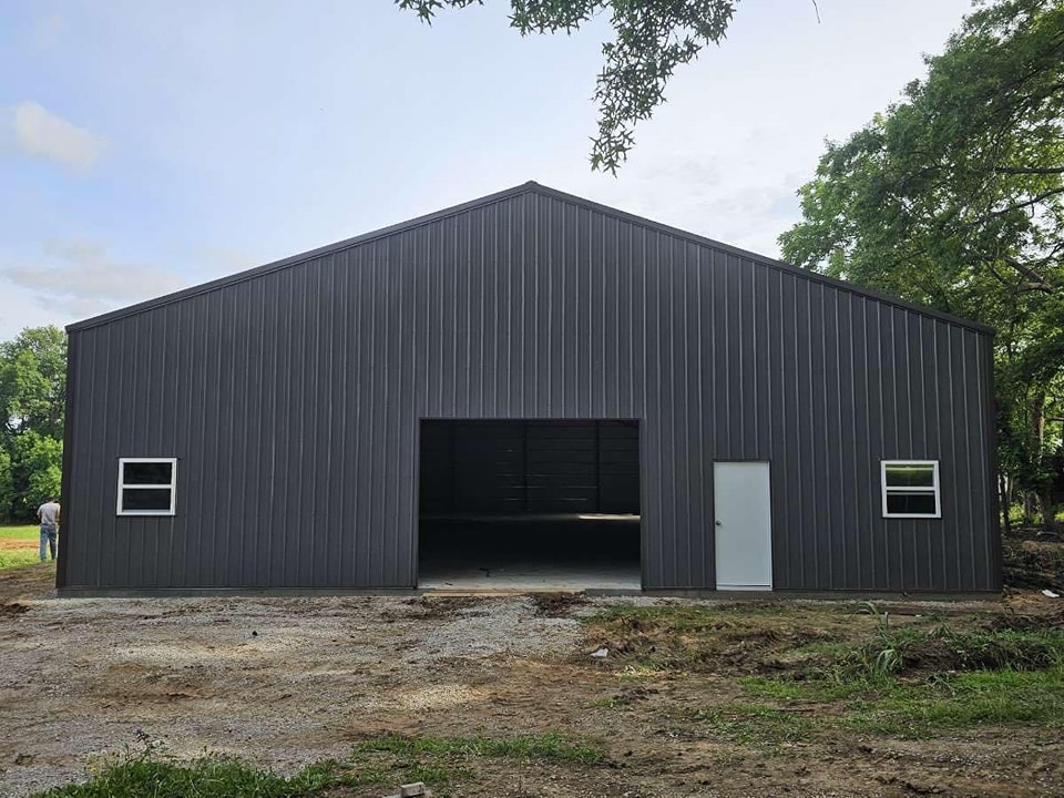 A large, newly constructed dark grey metal building with a high gable roof stands in an open area. It has a central large open space for vehicle entry flanked by two small windows and a white door on the right. The surrounding area is made of dirt and gravel.
