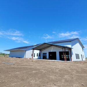 Large warehouse under clear blue sky, featuring metal siding and an extended covered entrance. The building is surrounded by a dirt lot, with construction materials visible nearby. No vegetation is present in the immediate area.