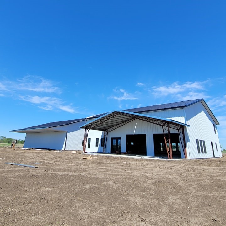 Large warehouse under clear blue sky, featuring metal siding and an extended covered entrance. The building is surrounded by a dirt lot, with construction materials visible nearby. No vegetation is present in the immediate area.