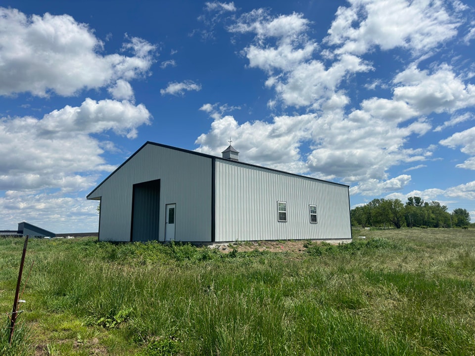 A large white metal barn stands in an open, grassy field under a partly cloudy sky. The barn features a peaked roof with a small cupola on top. The surrounding area is green, with trees visible in the distance.