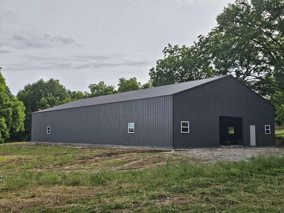A large, dark-gray metal building stands amid greenery and trees. The structure has a sloped roof, a few small windows, and a white door. The foreground shows a grassy area with patches of dirt and gravel, leading up to the building.