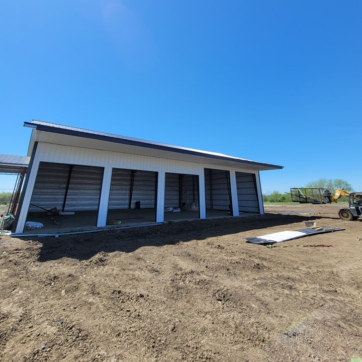 A newly constructed metal building with large open bays, set on a dirt lot. The sky is clear and bright, and construction materials are scattered around. A vehicle is visible near the structure.