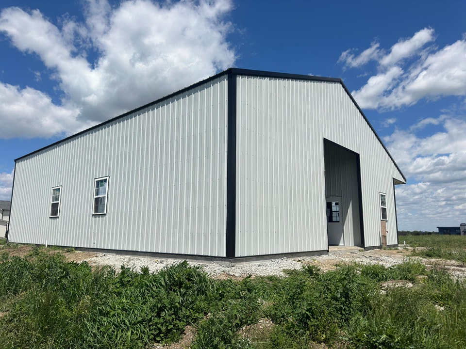 A large white metal building with a black roof stands on a gravel foundation surrounded by grass and wild plants. The structure has a high entrance and several windows. The sky is clear with a few clouds.