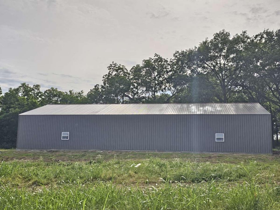 A large, gray, metal-sided building stands in a grassy area with trees lining the background. The building has a long, rectangular shape with a gently sloping roof. Two small square windows are visible on the side facing the viewer. The sky is overcast.