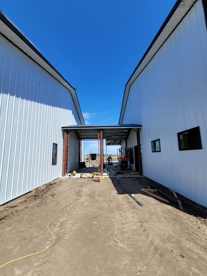 A view of a building under construction, with two white metal structures on either side and a connecting section in the middle. The ground is dirt, and construction materials are scattered around. A person is working in the background.