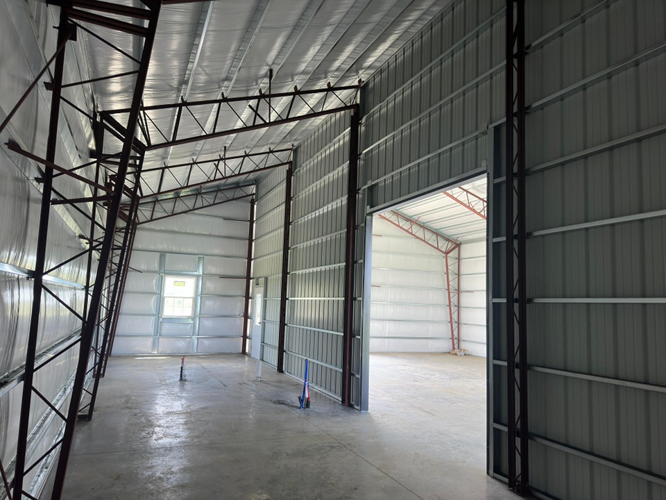 The image shows the interior of a large metal warehouse under construction. The space is open with high ceilings, exposed steel beams, and partially finished walls. Sunlight enters through a few openings, illuminating the concrete floor.