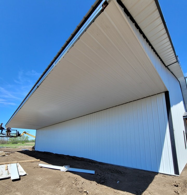 The image shows a large white metal structure with an open slanted roof, set against a clear blue sky. Several construction workers can be seen on the left side, working on the building. The ground is bare, with building materials scattered around.