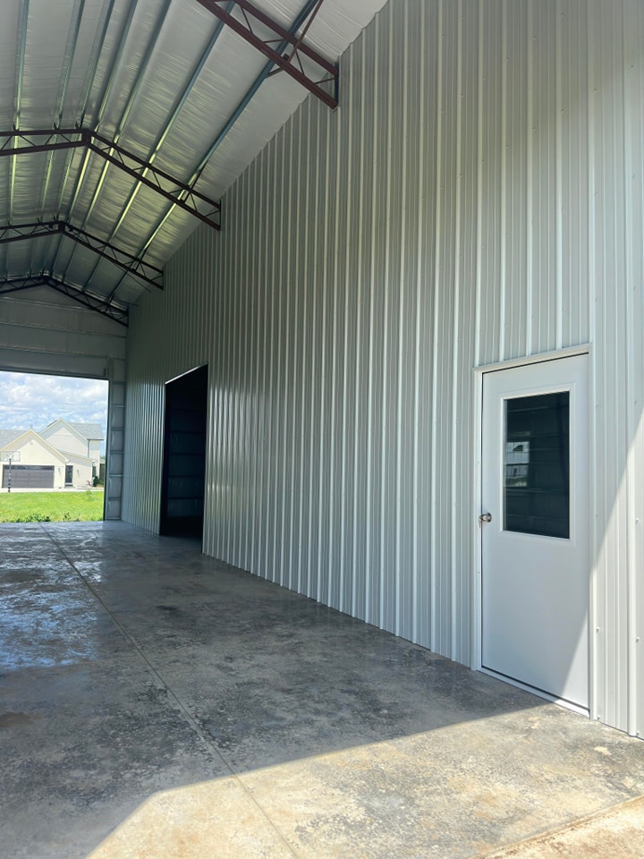 A large, spacious metal structure with a high ceiling, featuring a small white door with a window on the right side and a large open doorway on the left. The interior has a concrete floor. A small, light-colored house is visible in the background through the doorway.