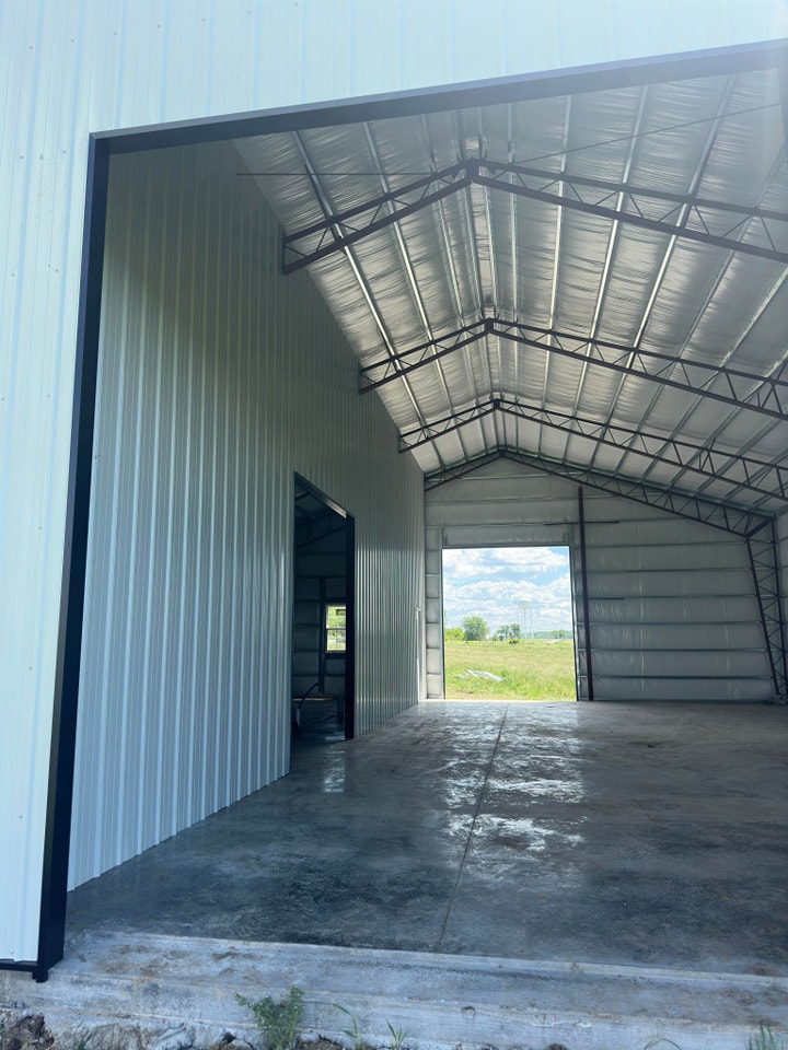 The interior of a large metal-framed building with an open-ended entrance. The walls and roof are constructed of corrugated metal. A clear open space leads to an outside view, showing a green field and a partly cloudy sky.