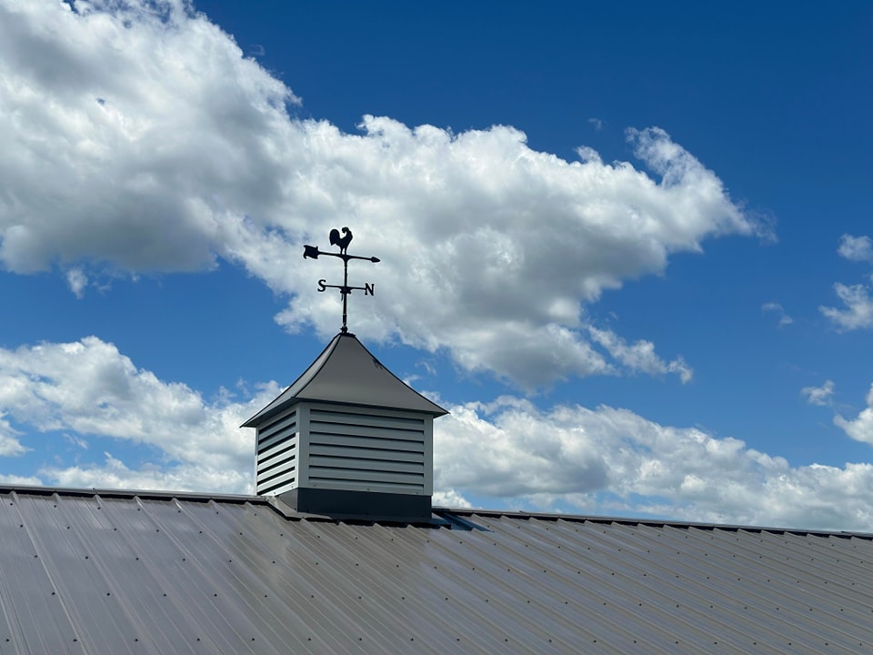 A rooftop with a metal weather vane shaped like a rooster, indicating north and south directions. The roof is slanted and made of metallic material. The sky above is blue with scattered, fluffy white clouds.