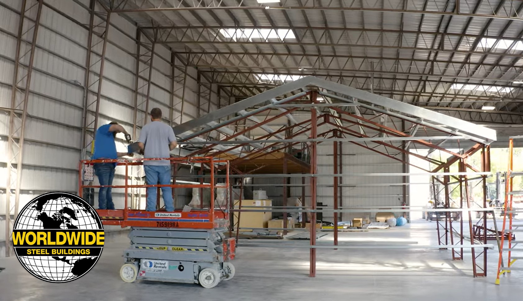 Two people standing on a scissor lift work on constructing a steel building framework inside a large warehouse. A "Worldwide Steel Buildings" logo is visible on the left side of the image. The open framework and high ceiling are prominently featured.