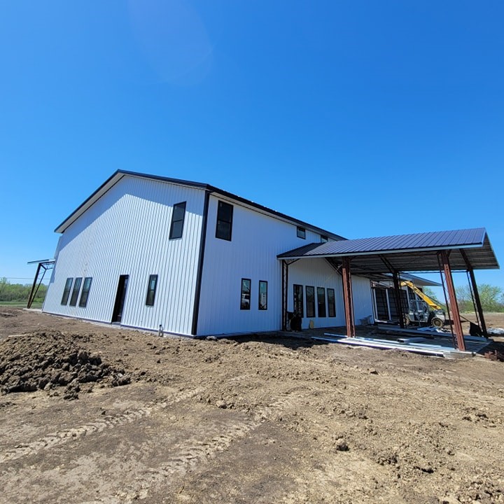 A two-story, white metal building under construction with a large covered entryway. Several windows line the sides, and a construction vehicle is visible nearby. The ground is dirt, and the sky is clear and blue.