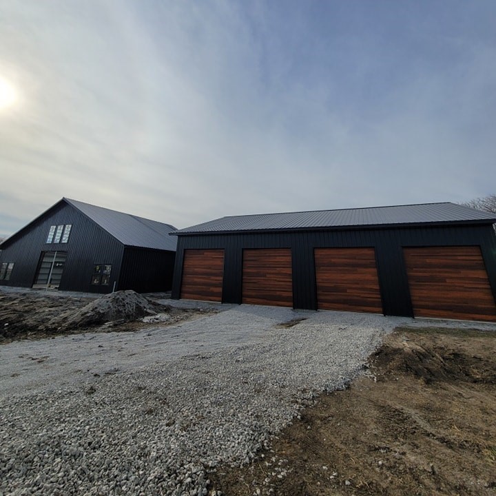 Modern black residential building with wood garage doors, peaked roof, and gravel driveway under clouds.