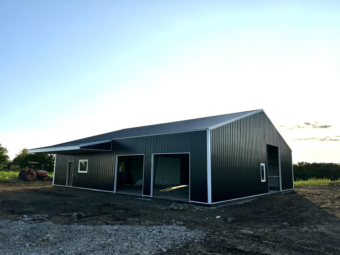 Barndominium with metal roof on gravel lot, red tractor nearby, trees and clear sky in background.