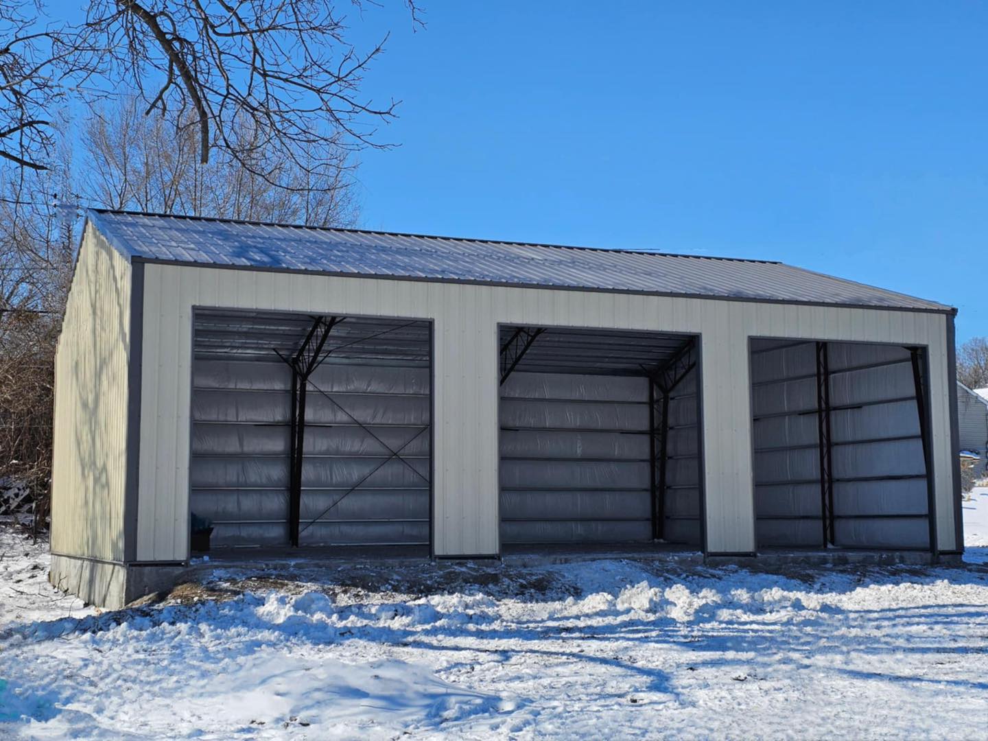 Metal barn with open entrances on snowy ground, clear blue sky, and bare trees in the background.