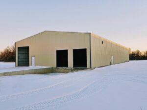 A large beige warehouse structure stands in a snowy field, showcasing ACCI's specialty materials. The building has three open entrances on one side and is surrounded by snow-covered ground under a clear sky, with tracks etched in the snow leading to the impressive structure.