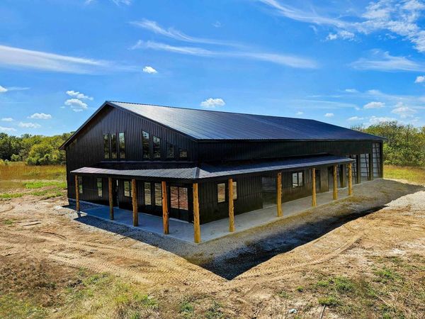 Black barndominium with gabled roof and many windows in an open field under a blue, cloudy sky.