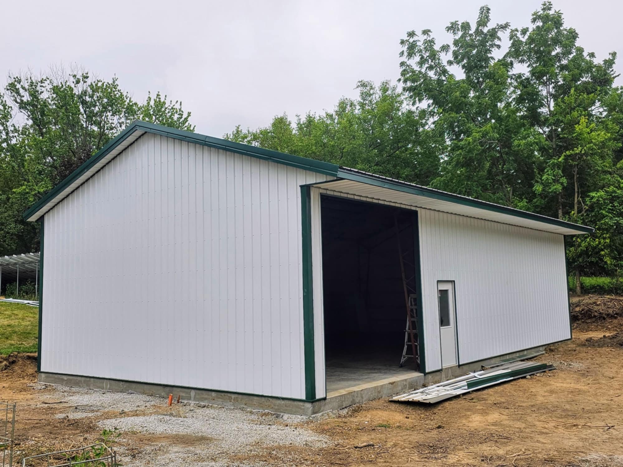 White building with green roof on gravel, open doorway, trees; future development site.