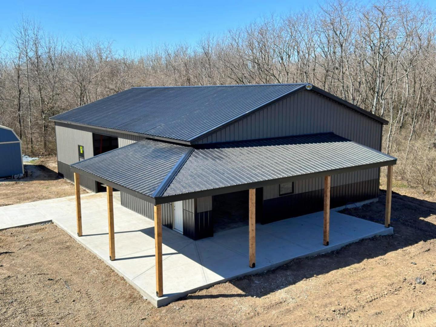 Gray single-story building with black roof and porch, near leafless trees and smaller structure.