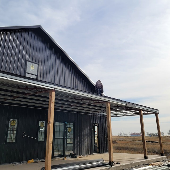 Person on roof of dark house under construction, wooden pillars, overlooks open land and clear sky.