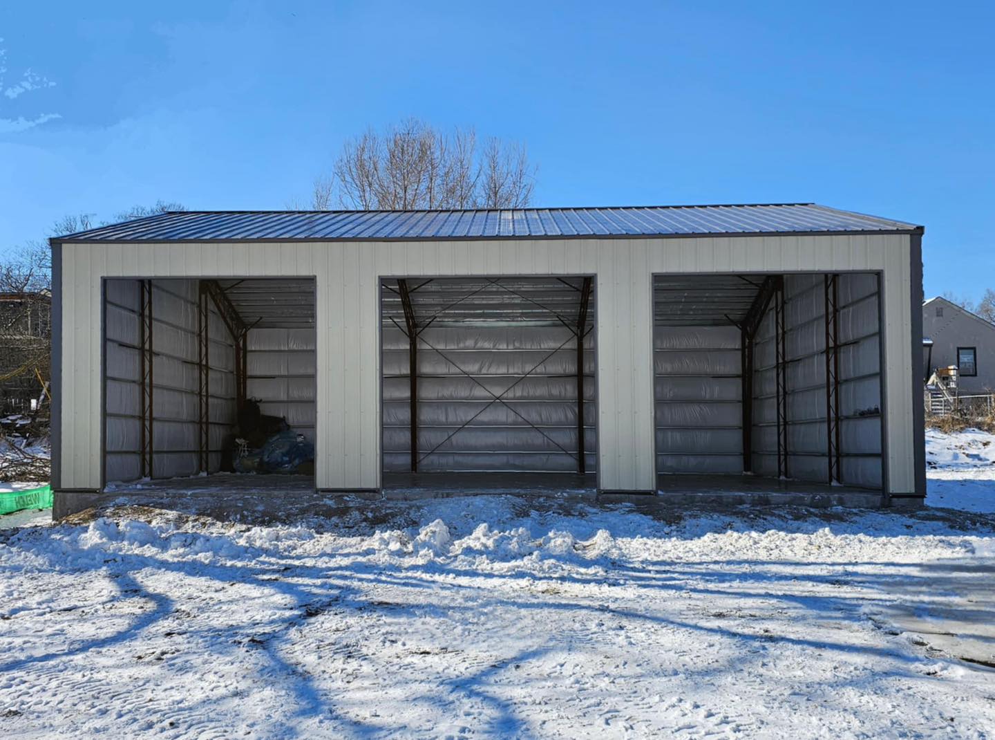 Metal garage with three open bays on snowy ground; gray exterior, trees, blue sky backdrop.