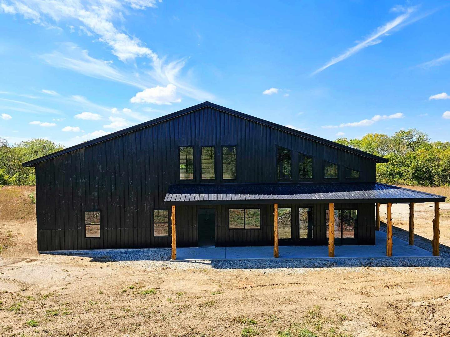 Black barndominium with tall windows and overhang on dirt lot under blue sky with scattered clouds.